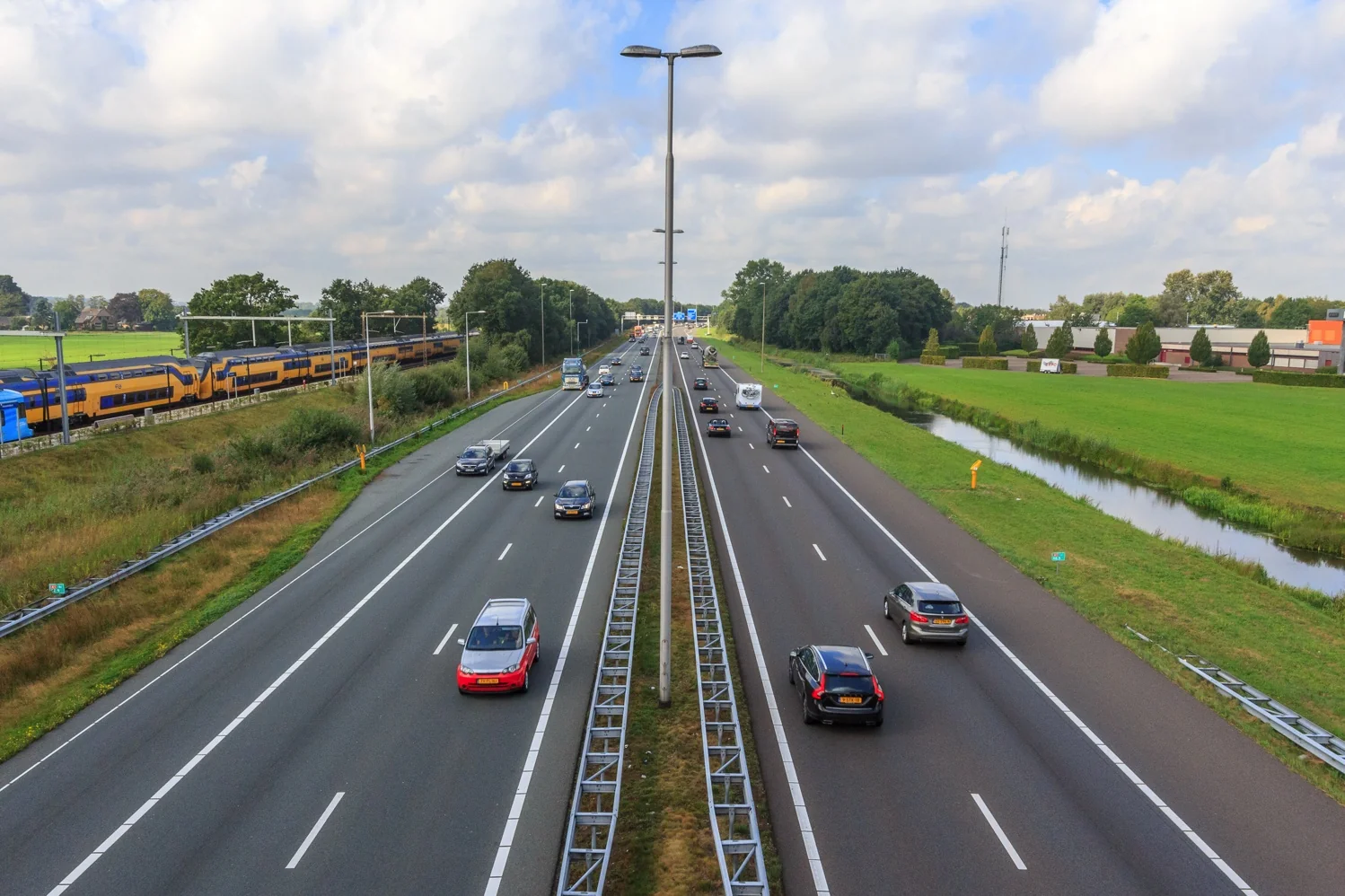 Drukke snelweg met naastgelegen spoorlijn waarop een NS-trein rijdt, omgeven door groen landschap bij De Wel.